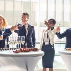 Asistentes brindan durante un evento tipo cóctel con mesas altas, bebidas y aperitivos.